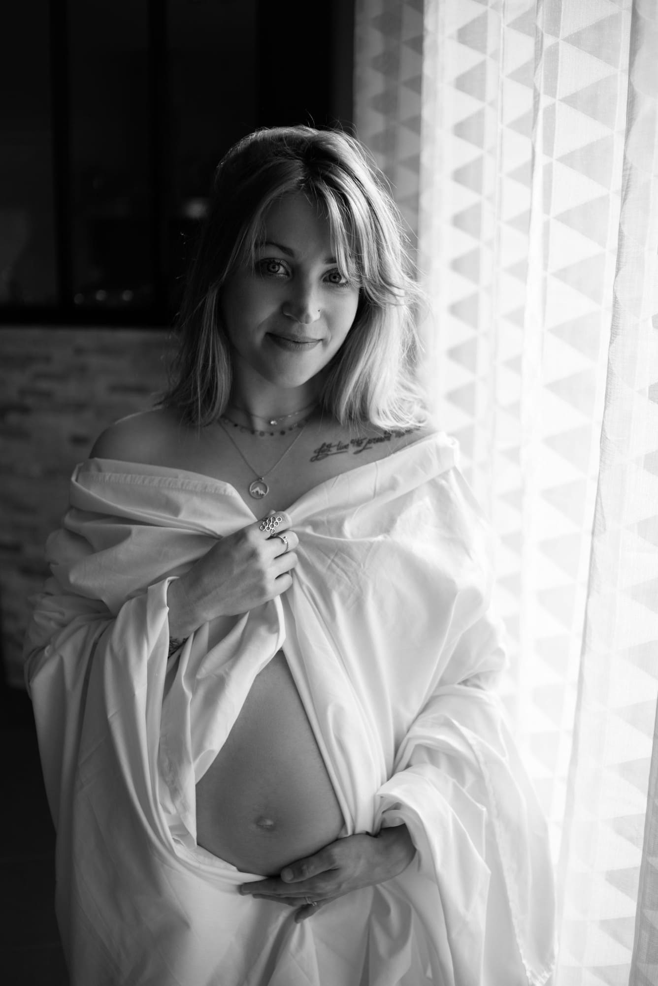 La photo est en noir et banc. On voit une jeune femme vêtue d'une chemise blanche, ouverte sur son ventre rond de femme enceinte.